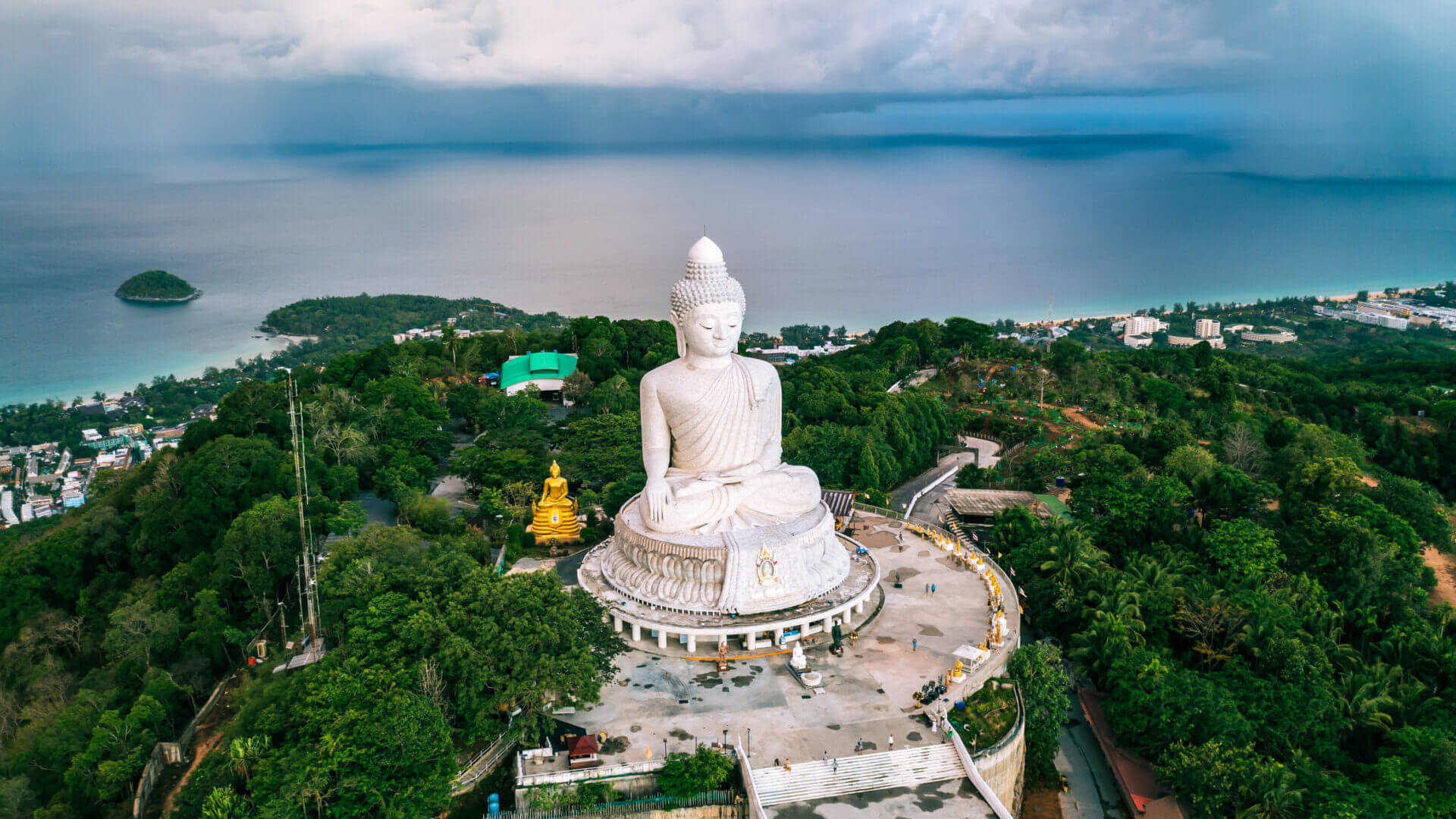A large white Buddha statue overlooking a green forest and water body.